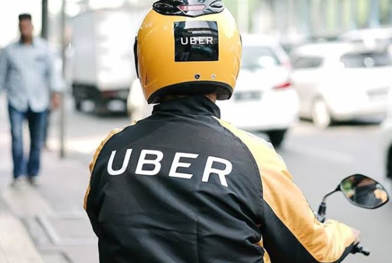Motoqueiro com capacete amarelo e com a marca da Uber aparecendo na parte de trás. O nome da uber também aparece na jaqueta. Ao fundo, uma pessoa caminha e carros passam.
