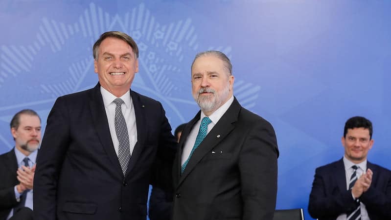 Jair Bolsonaro posa para foto ao lado de Augusto Aras. Bolsonaro está sorrindo de forma expansiva. Ao fundo, Sérgio moro aparece sentado, aplaudindo, e olhando para Aras e Bolsonaro.