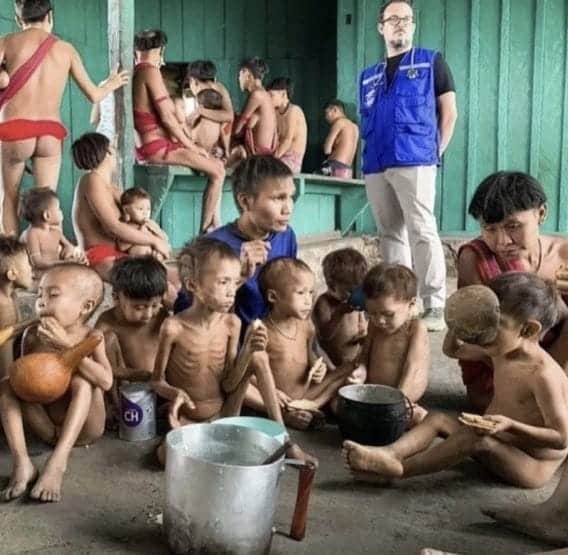 Crianças aparecem desnutridas diante de um caldeirão de alumínio. Elas estão sentadas e, ao fundo, um agente do SUS aparece em pé