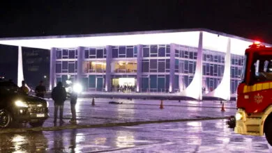 Foto mostra corpo caído no fundo da foto, diante do prédio do STS. Policiais estão conversando no canto esquerdo da imagem.